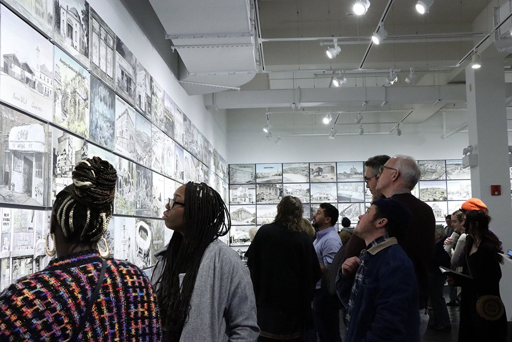 A group of people stand in the art gallery, looking and pointing at a black and white display.