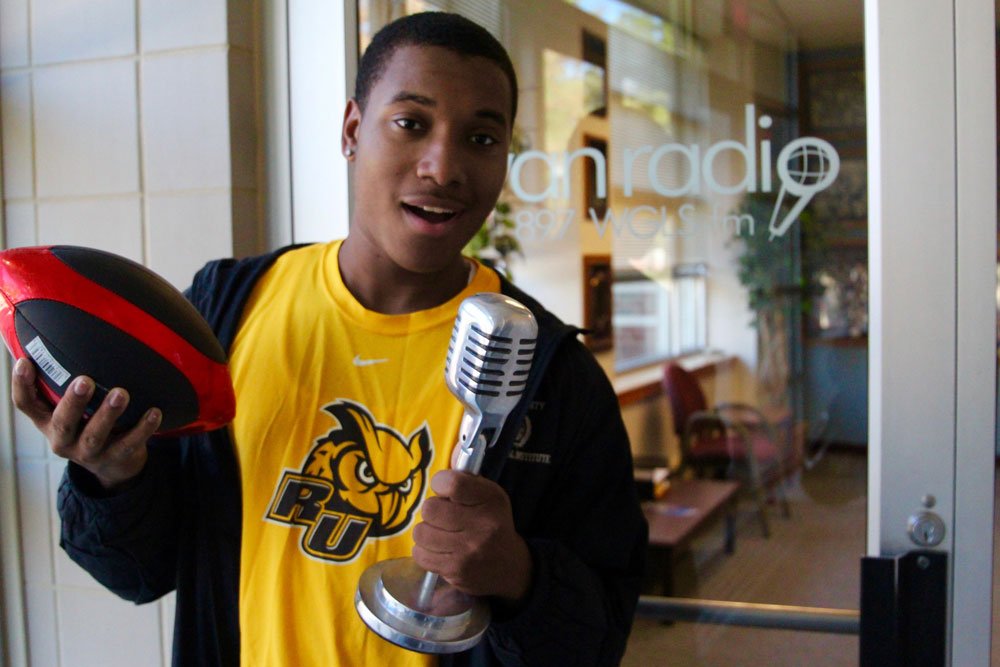student stands outside of radio station window holding a football and a microphone as if he's talking into it