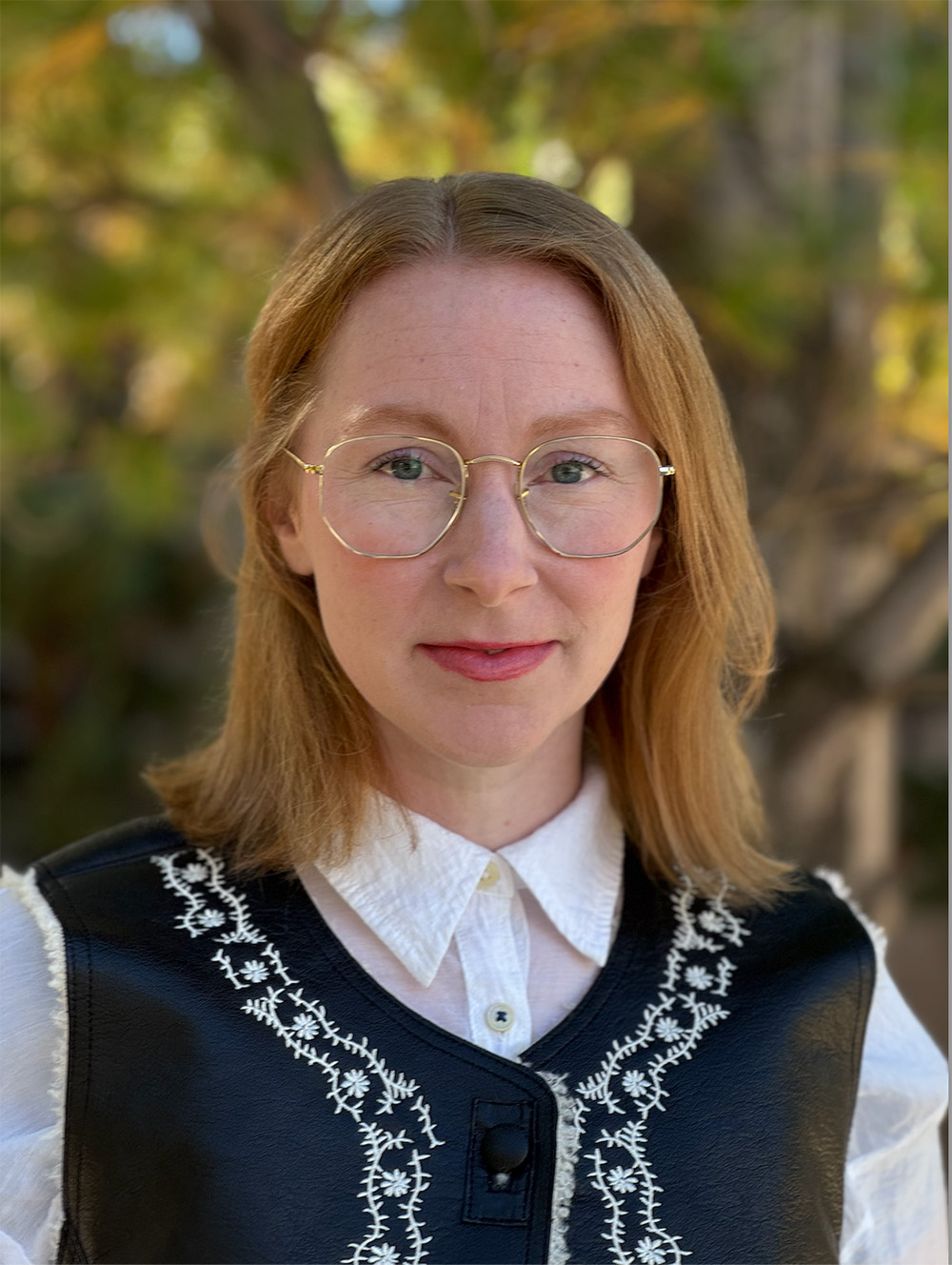 Audrey Ellis wearing glasses, a white shirt and embellished black vest, smiling at the camera.