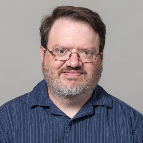 Andy Carlson wearing a blue striped shirt, glasses and a beard, smiling at the camera. 