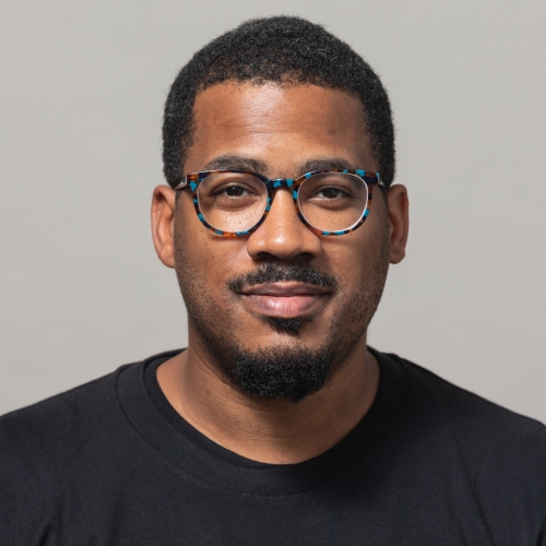 James Lewis wearing a black shirt and round blue glasses, smiling at the camera.