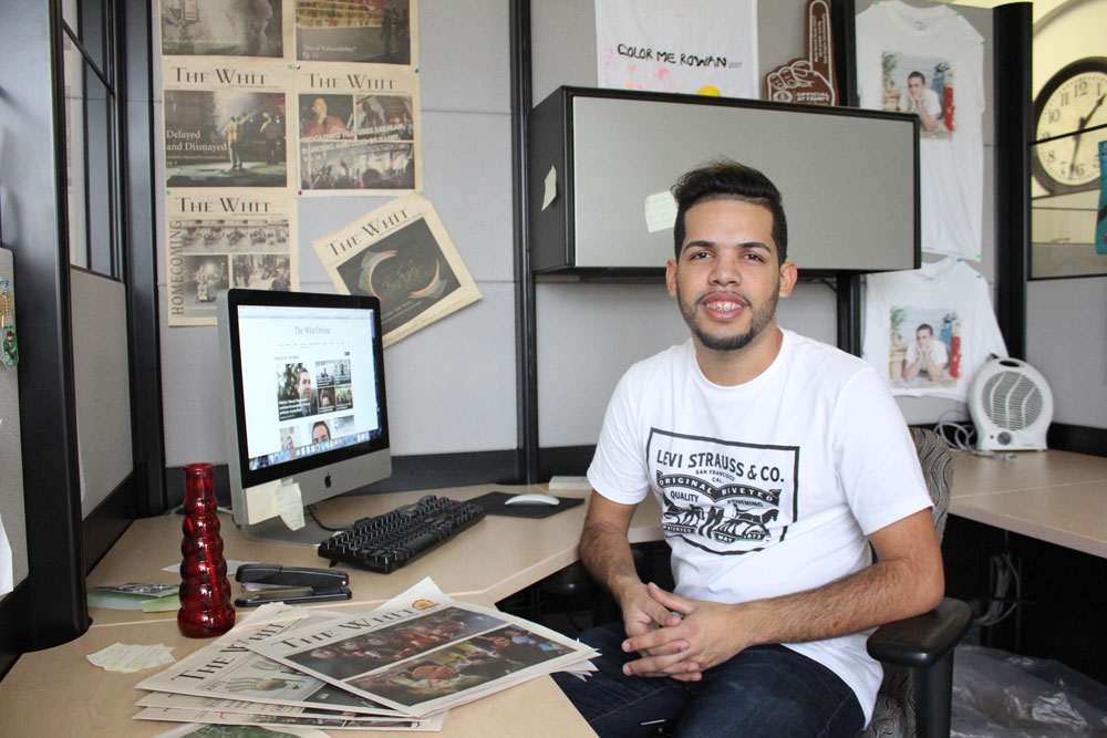 male student sits at a desk in the student newspaper room, surrounded by work